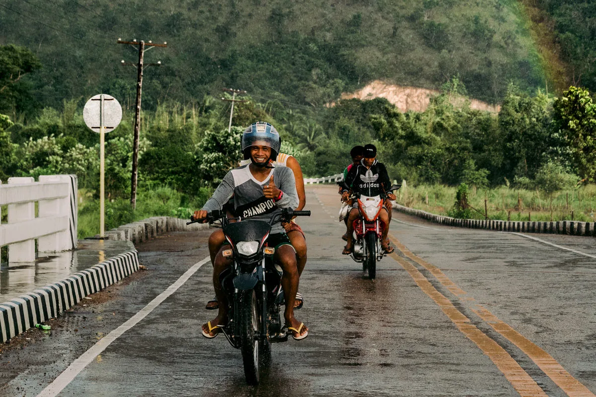 Smiling men riding motorbikes along a wet road past green hills, with a faint rainbow above, in Coron, Palawan, Philippines