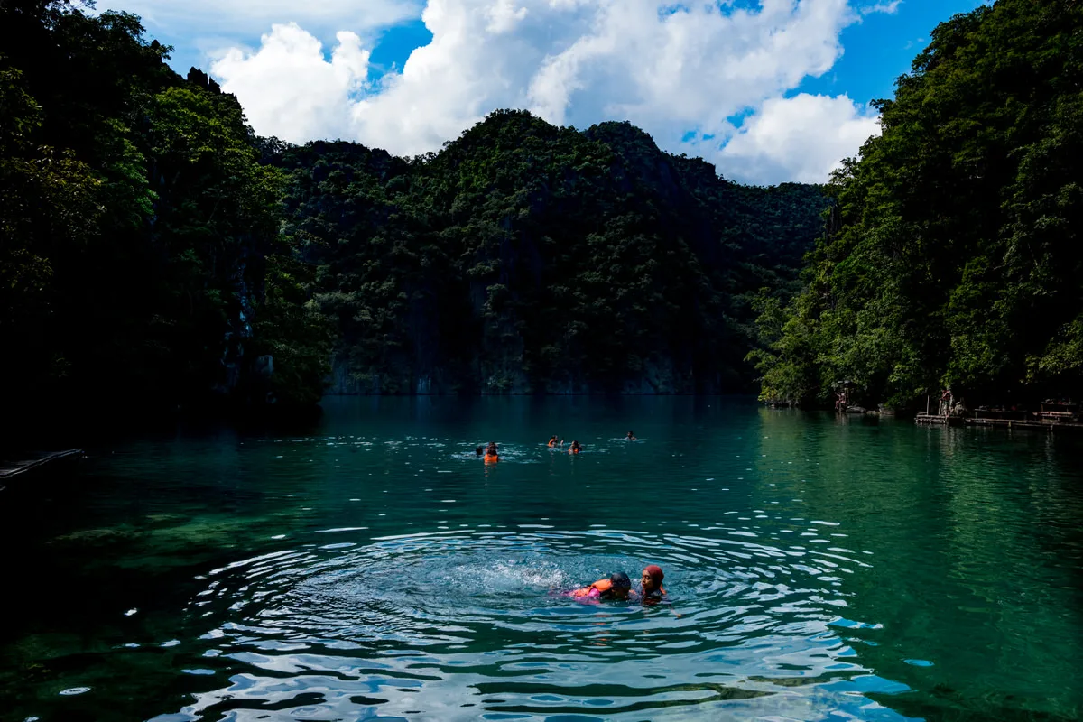 Swimmers in life jackets floating in a clear green lagoon surrounded by steep forested cliffs in Coron, Palawan, Philippines