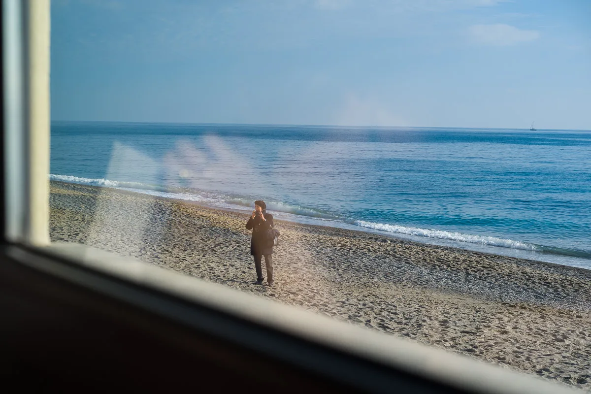 Person photographing the sea from the beach, seen through the restaurant window of Osteria Rivamare in Borgio Verezzi, Italy