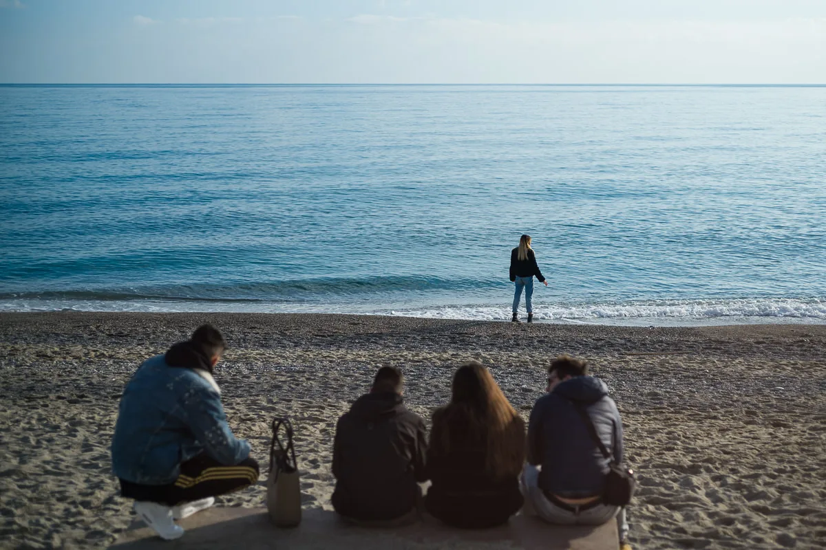 Four friends sitting on a ledge watching a woman at the water's edge on a sunny winter beach in Borgio Verezzi, Italy
