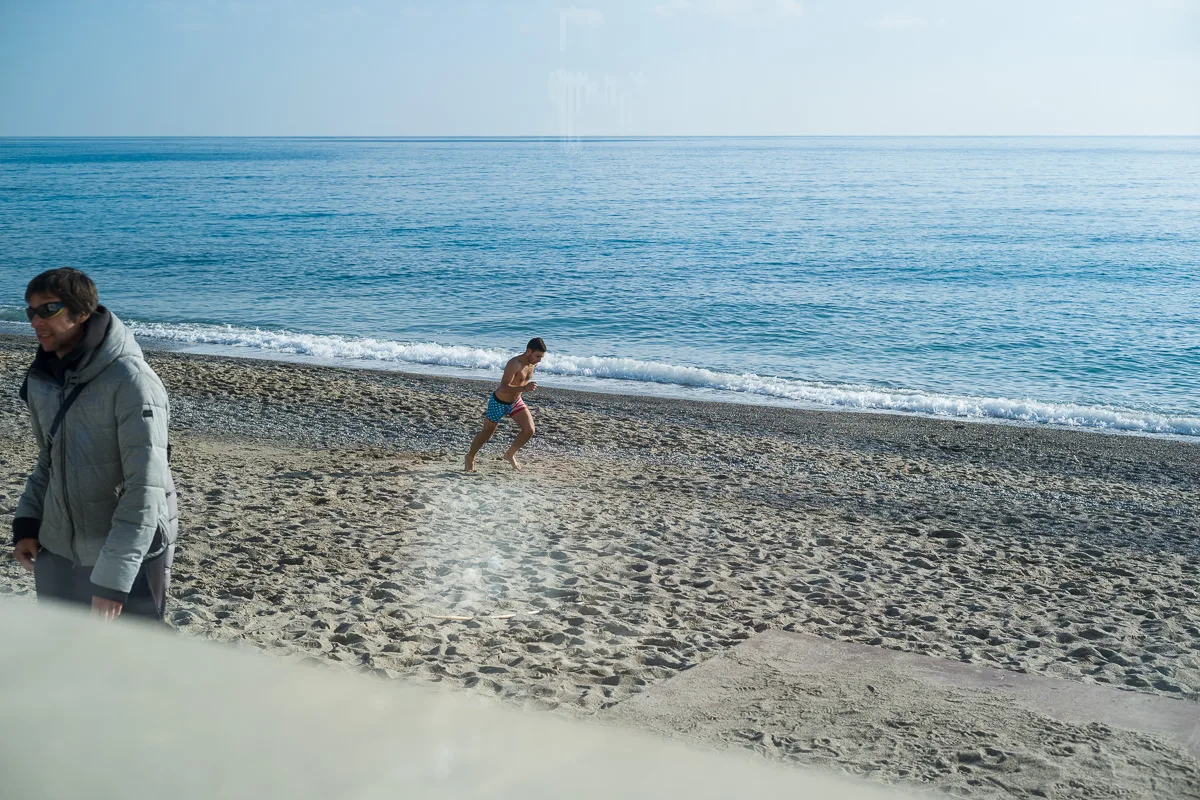 Swimmer in trunks running across the sand toward the sea while a man in a winter jacket walks by in Borgio Verezzi, Italy