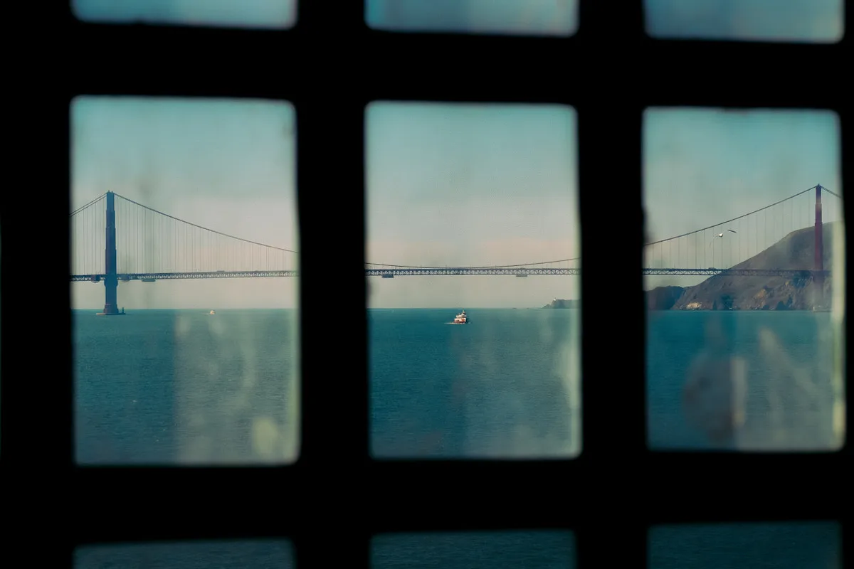 Golden Gate Bridge and a passing boat seen through the barred, weathered windows of a building on Alcatraz Island, San Francisco