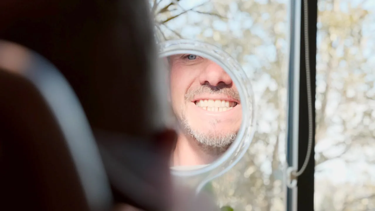 Still from a film for Peak Dental Health: a smiling man checking his teeth in a round hand mirror by a sunny window