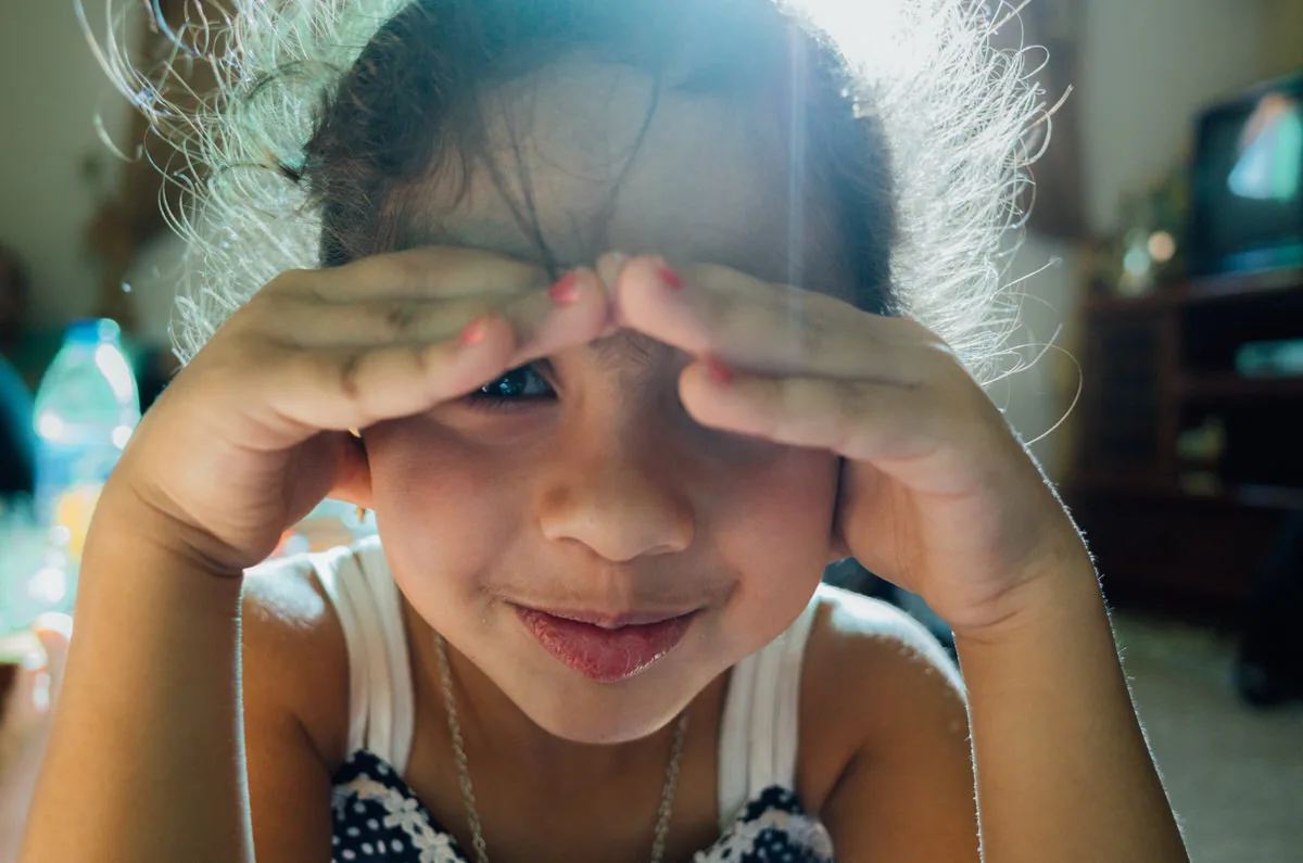 Young girl smiling as she peeks out from under her hands, backlit by sunlight streaming into a room