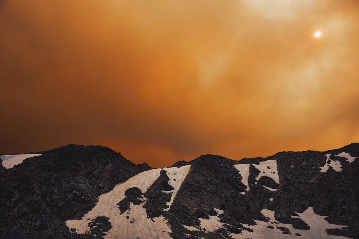 Orange wildfire smoke filling the sky and dimming the sun over dark, snow-streaked mountain ridges near Nederland, Colorado