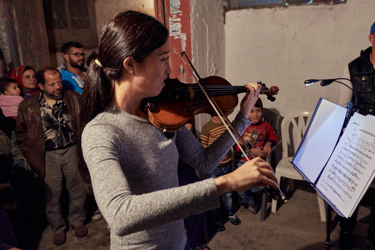 A violinist plays from sheet music in Said Gawash camp, Beirut, as children and families watch from the alley behind her