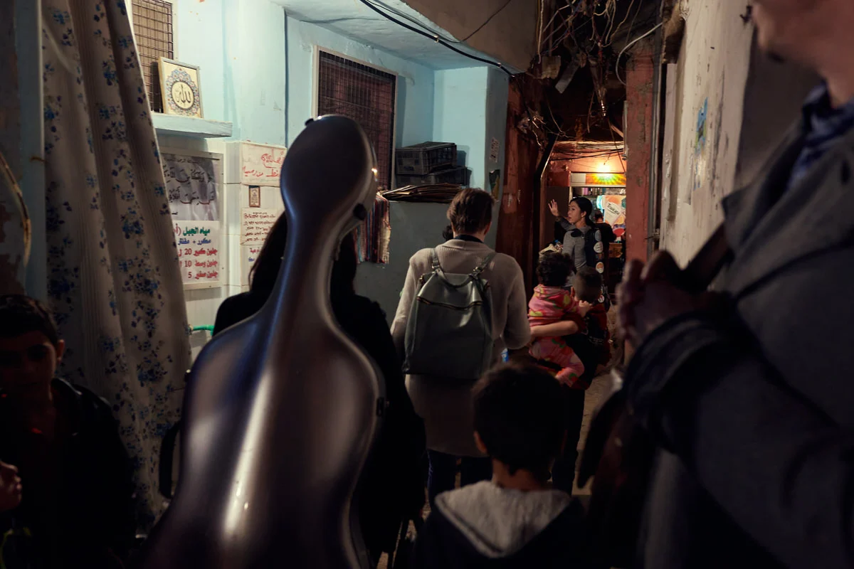 Musicians carry a cello case through a narrow alley of Said Gawash camp, Beirut, as a woman ahead raises her hand