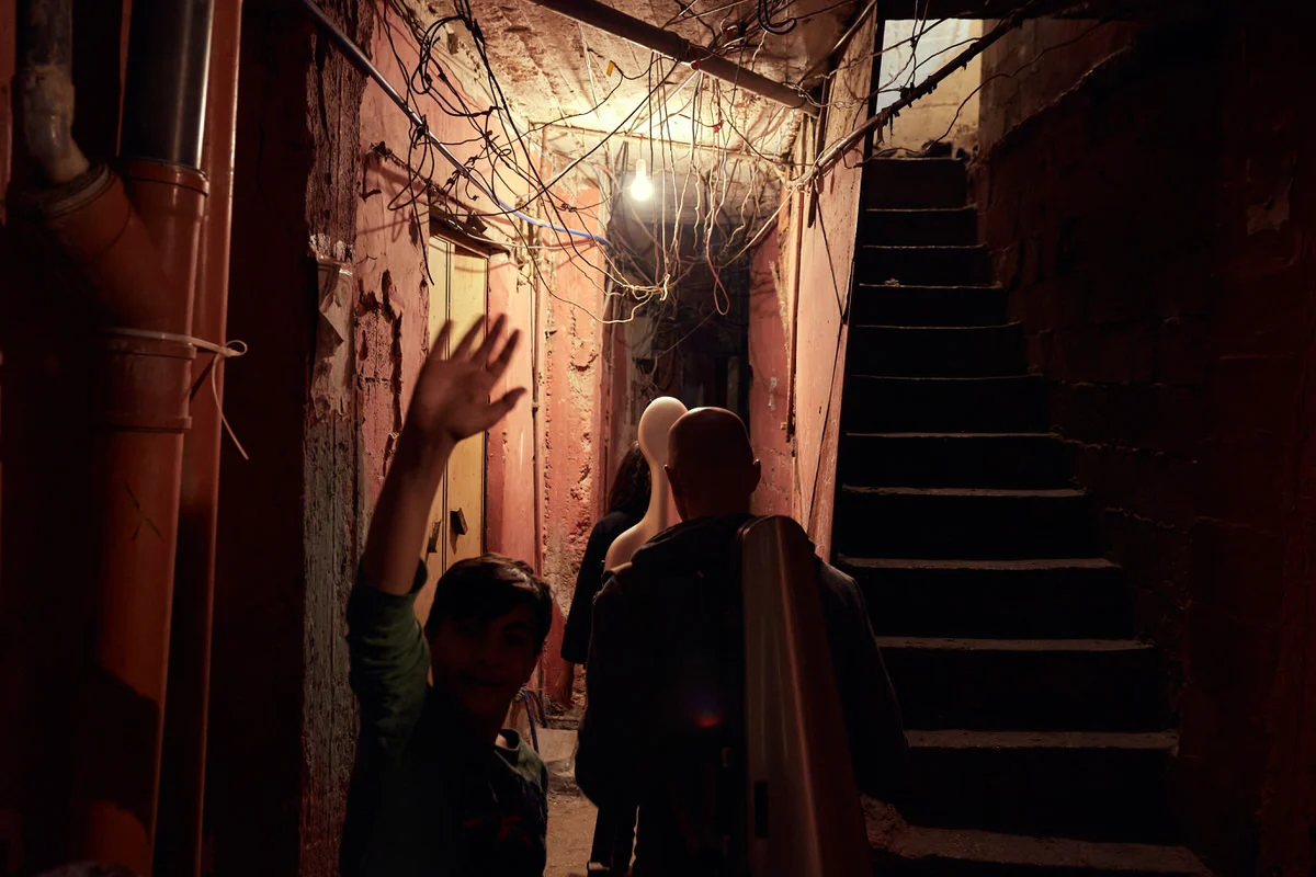 A boy waves as a man carrying a cello case walks a dim, wire-strung alley in Said Gawash camp, Beirut, past a mannequin