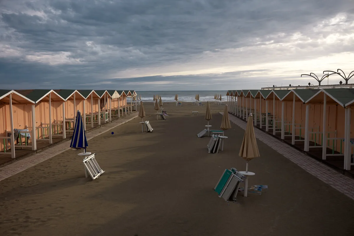 Empty beach with rows of wooden cabins, closed umbrellas and stacked loungers under a stormy sky in Ostia, Italy