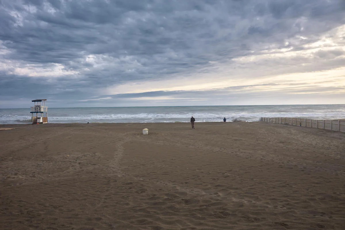 Wide empty beach with a lifeguard tower and two distant walkers by the surf under heavy gray clouds in Ostia, Italy