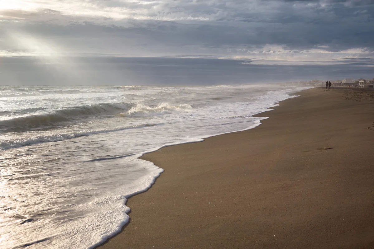 Waves washing onto a long empty beach as sunlight breaks through the clouds, with two small figures far away, in Ostia, Italy