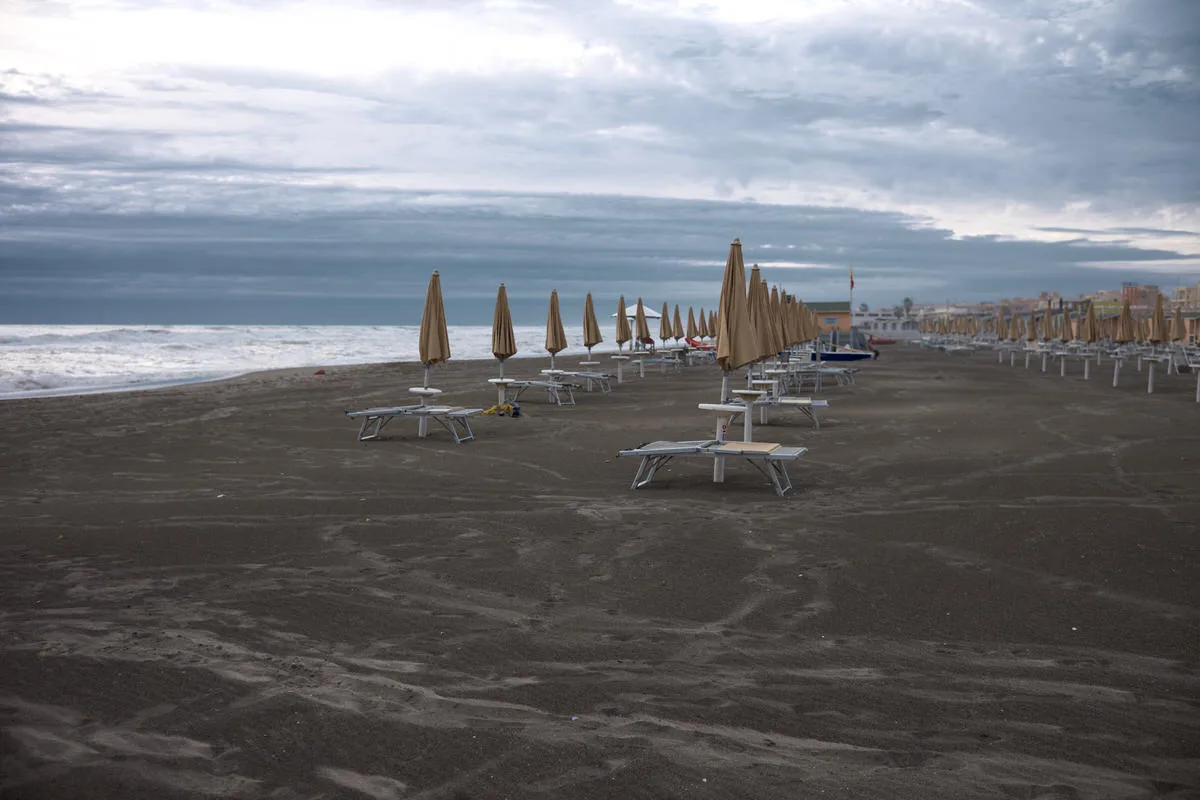 Rows of closed beach umbrellas and empty sun loungers on dark sand along the sea under a cloudy sky in Ostia, Italy