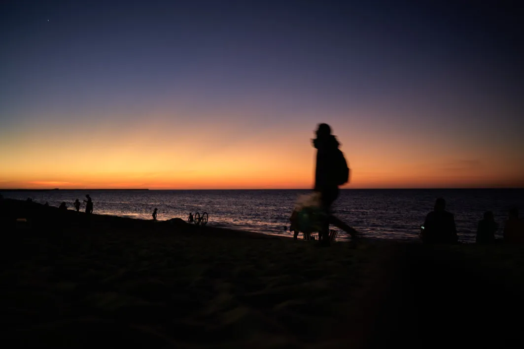 Silhouettes of people on the beach at dusk, with a blurred figure walking past an orange and violet sky over the sea in Fortaleza