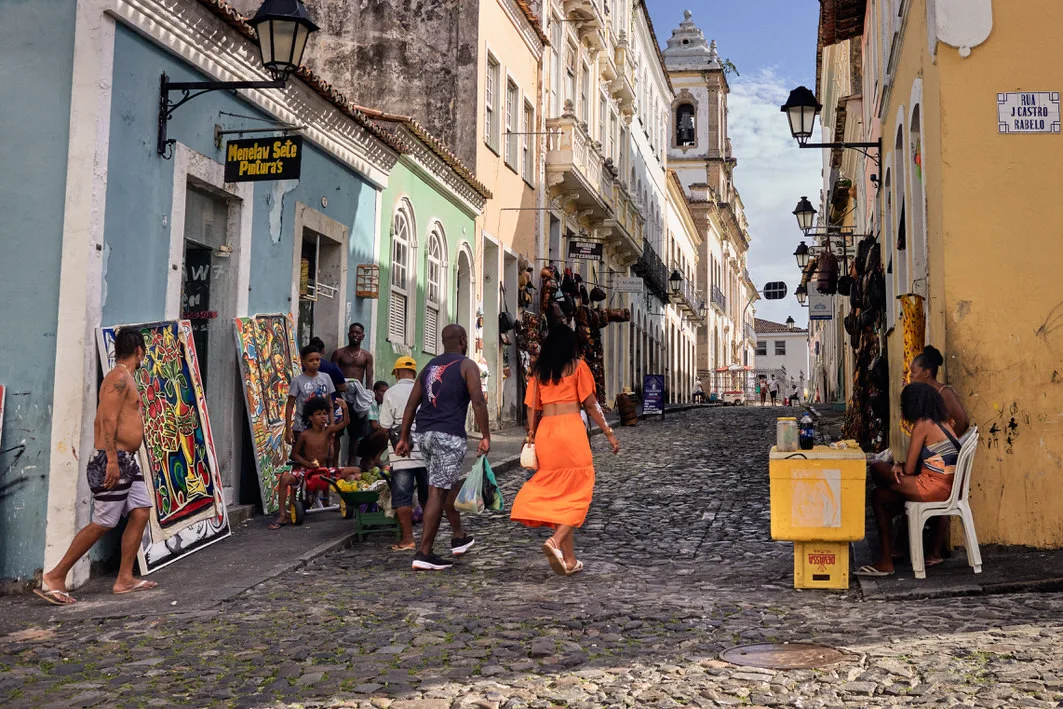 Woman in an orange dress crosses a cobbled street past painting sellers and vendors in Salvador's historic center