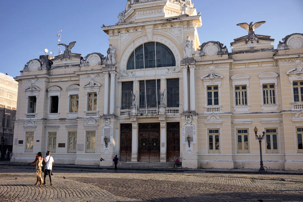 Couple crossing a cobbled square in front of the ornate Palácio Rio Branco, topped with eagle statues, in Salvador, Bahia