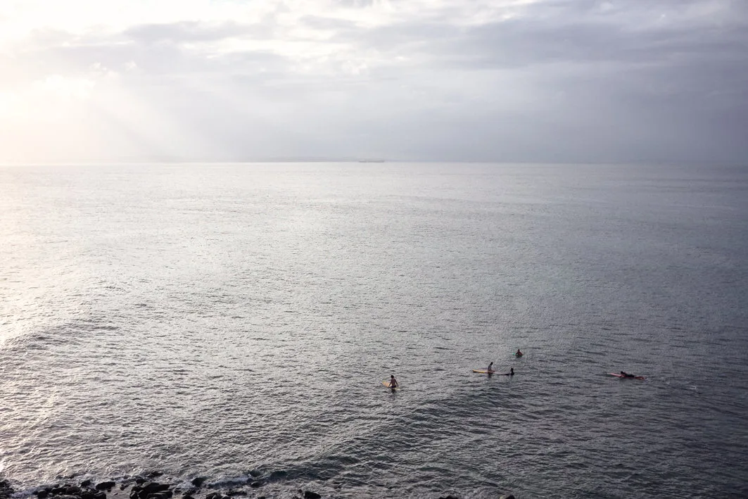 Surfers waiting on a calm silver sea under rays of light breaking through clouds, seen from Morro do Cristo, Salvador