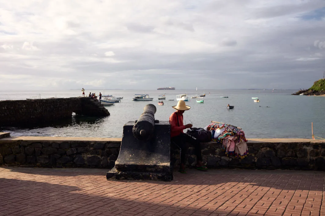 Vendor in a straw hat seated by an old cannon with a rack of colorful sarongs on the seawall at Porto da Barra, Salvador