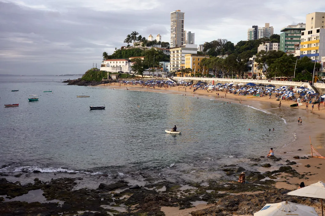 Crowded beach umbrellas, swimmers and small boats in the calm bay of Porto da Barra beneath hillside buildings, Salvador, Brazil