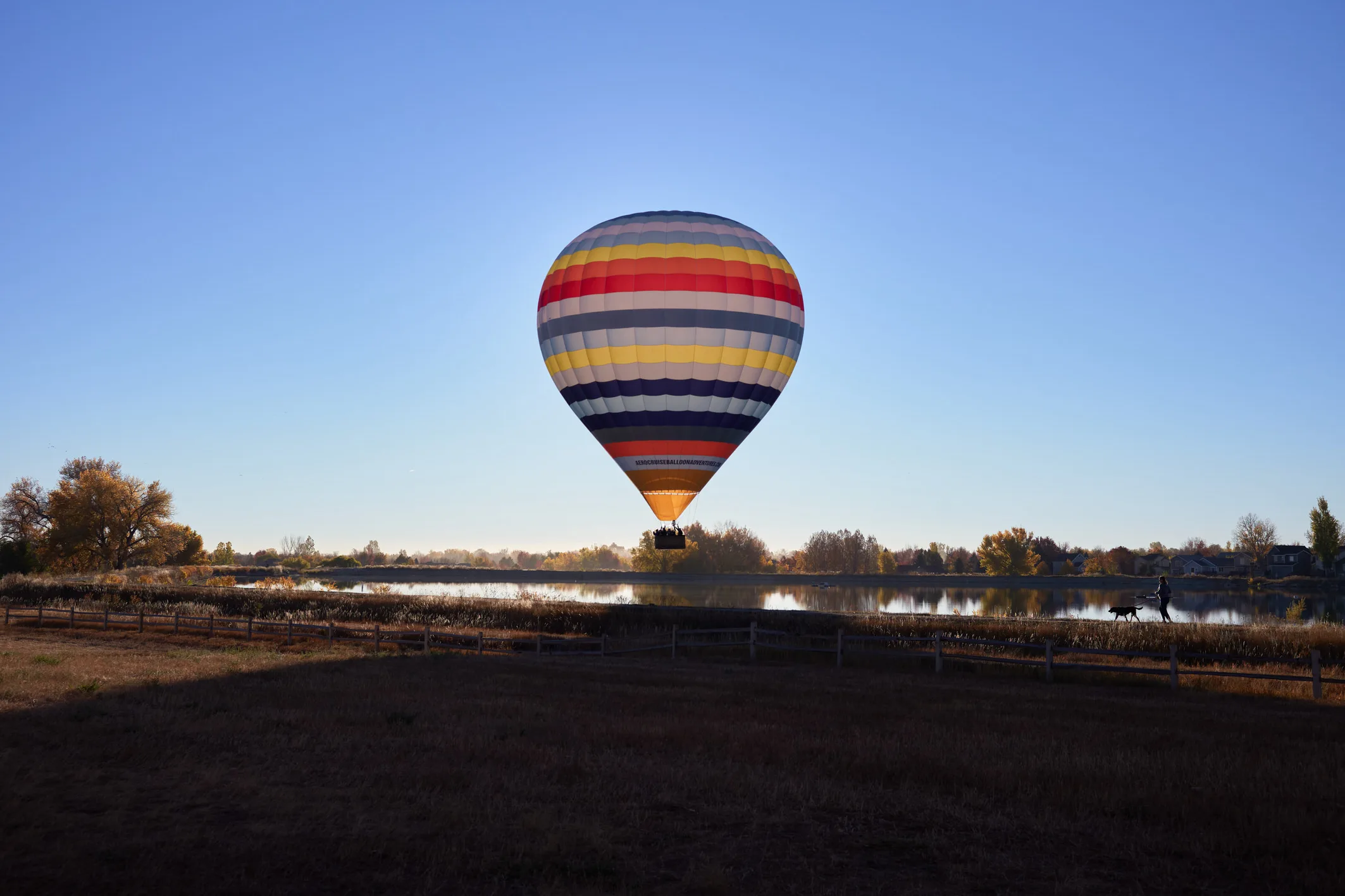Striped hot air balloon lifting off over a still lake at dawn while a person walks a dog along the autumn shore