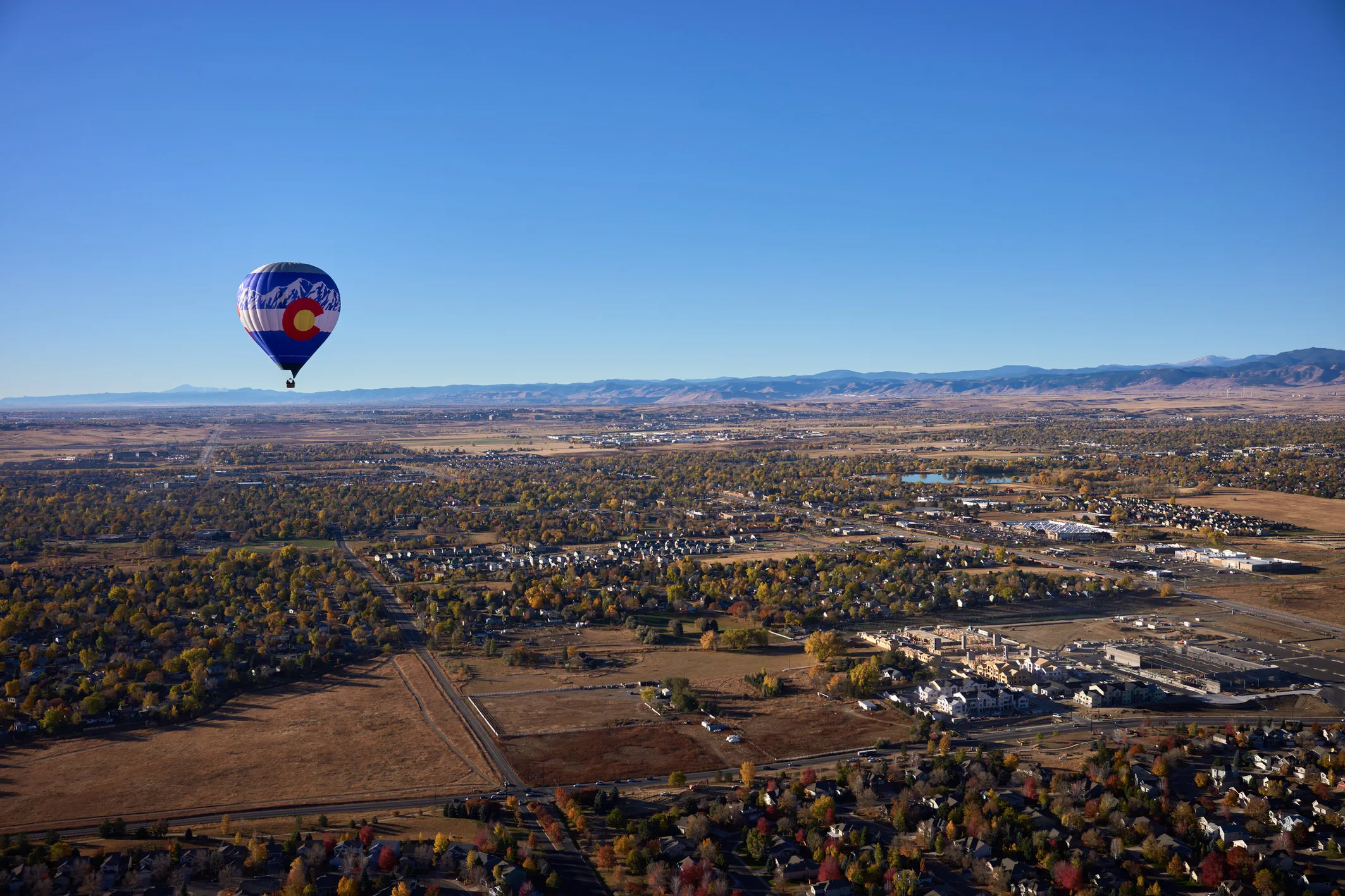 Balloon with the Colorado flag floating over autumn suburbs, with the Front Range mountains on the horizon