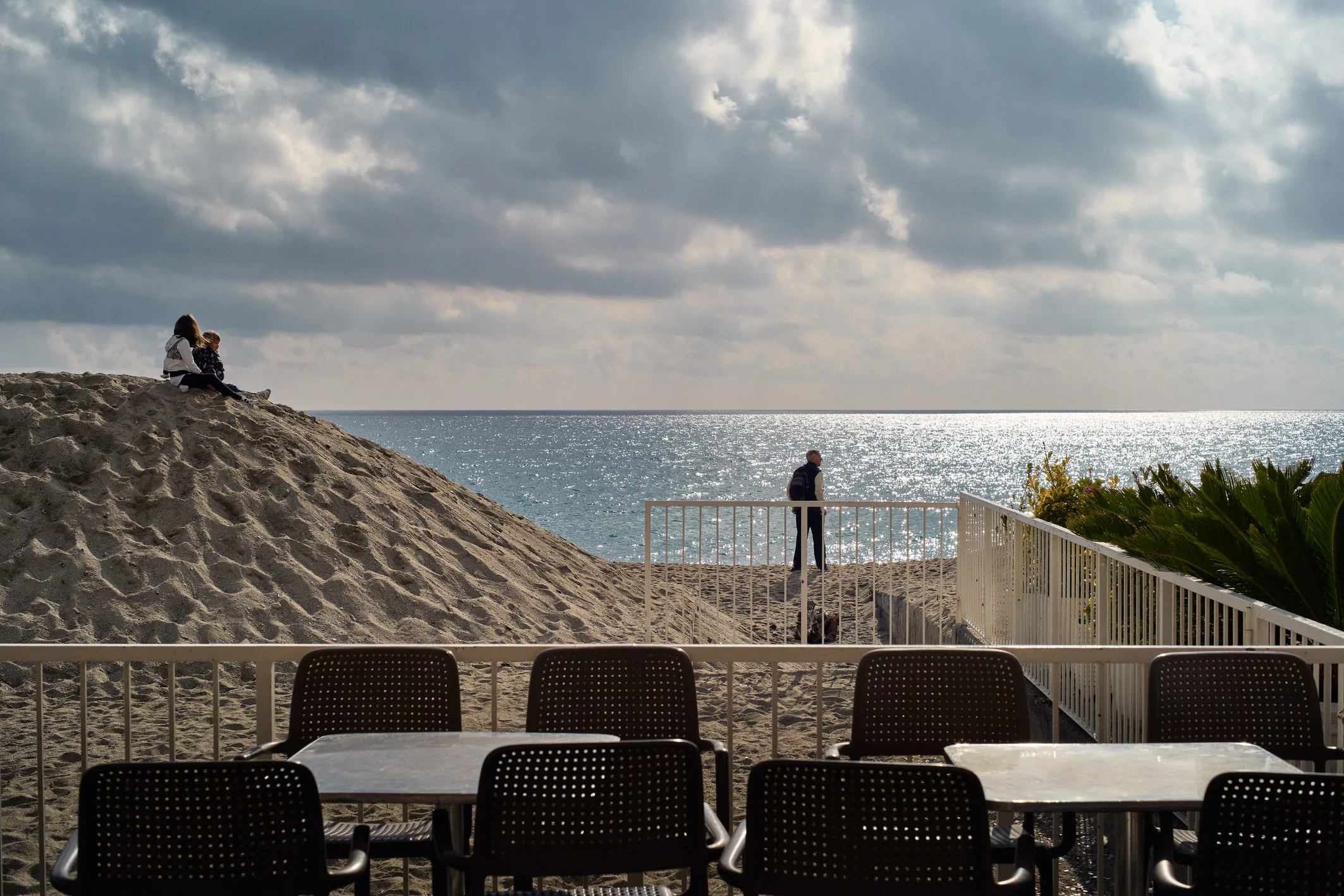 Empty café terrace tables facing a sand mound where two people sit above the glittering winter sea at Finale Ligure