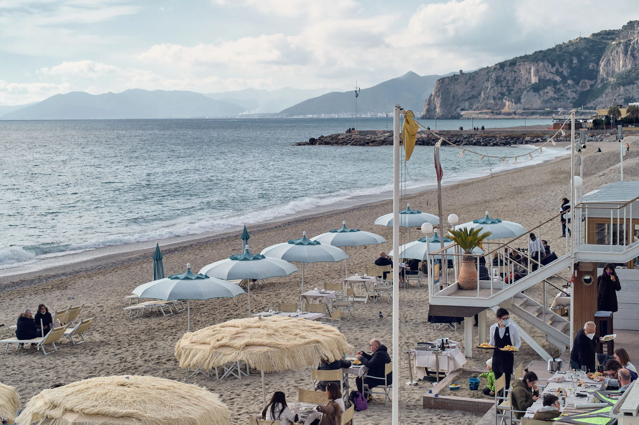 Waiters serving lunch at a beach restaurant under umbrellas on a mild winter day, cliffs behind, Finale Ligure