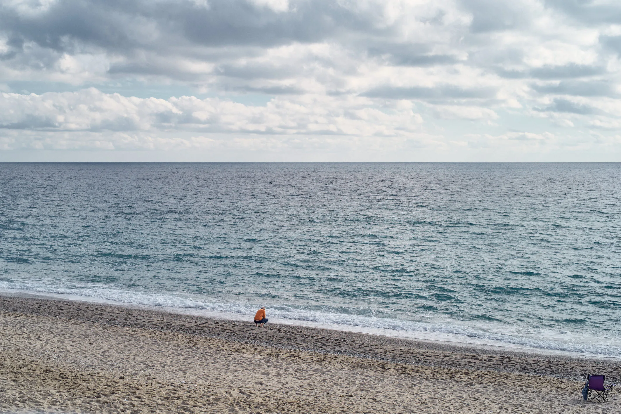 Lone figure in an orange jacket crouching at the water's edge on a wide winter beach under gray clouds, Finale Ligure, Italy