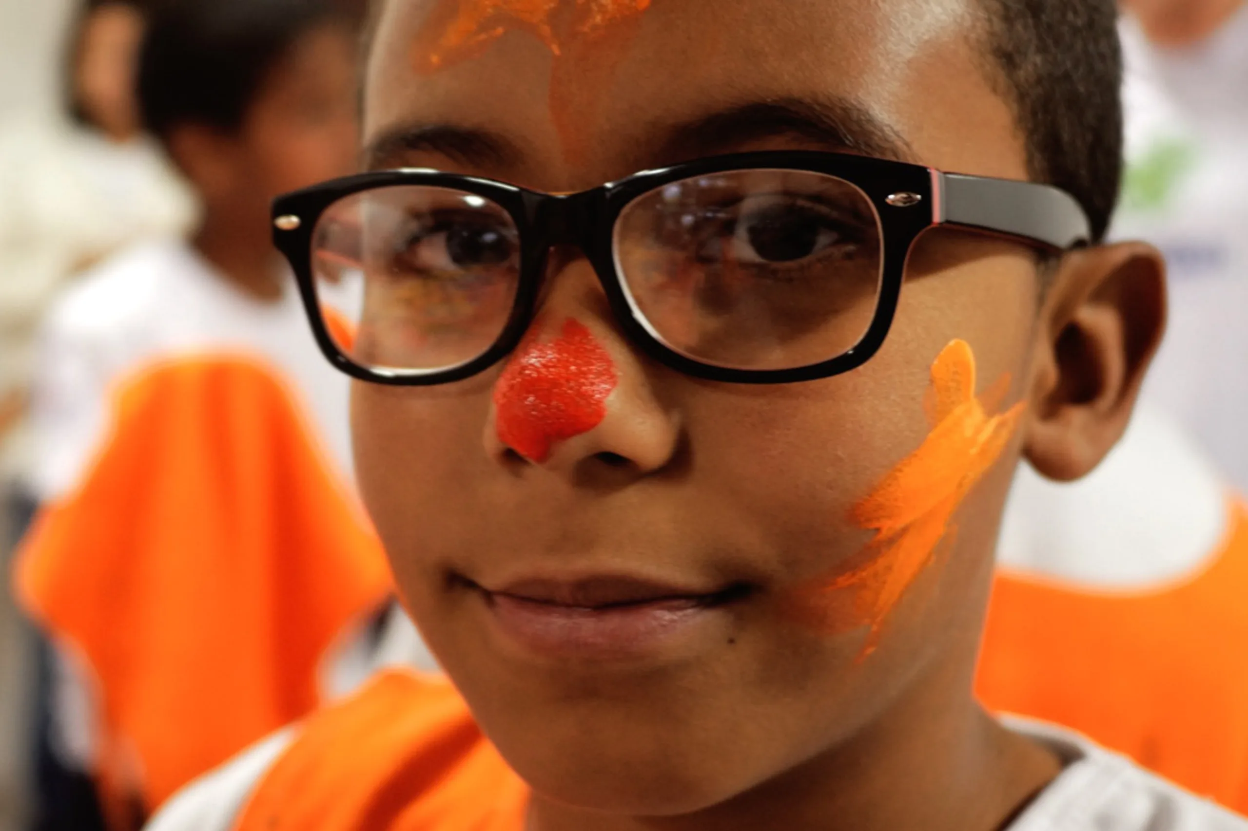 A boy in glasses with a painted red nose and orange face paint at a workshop, Belo Horizonte
