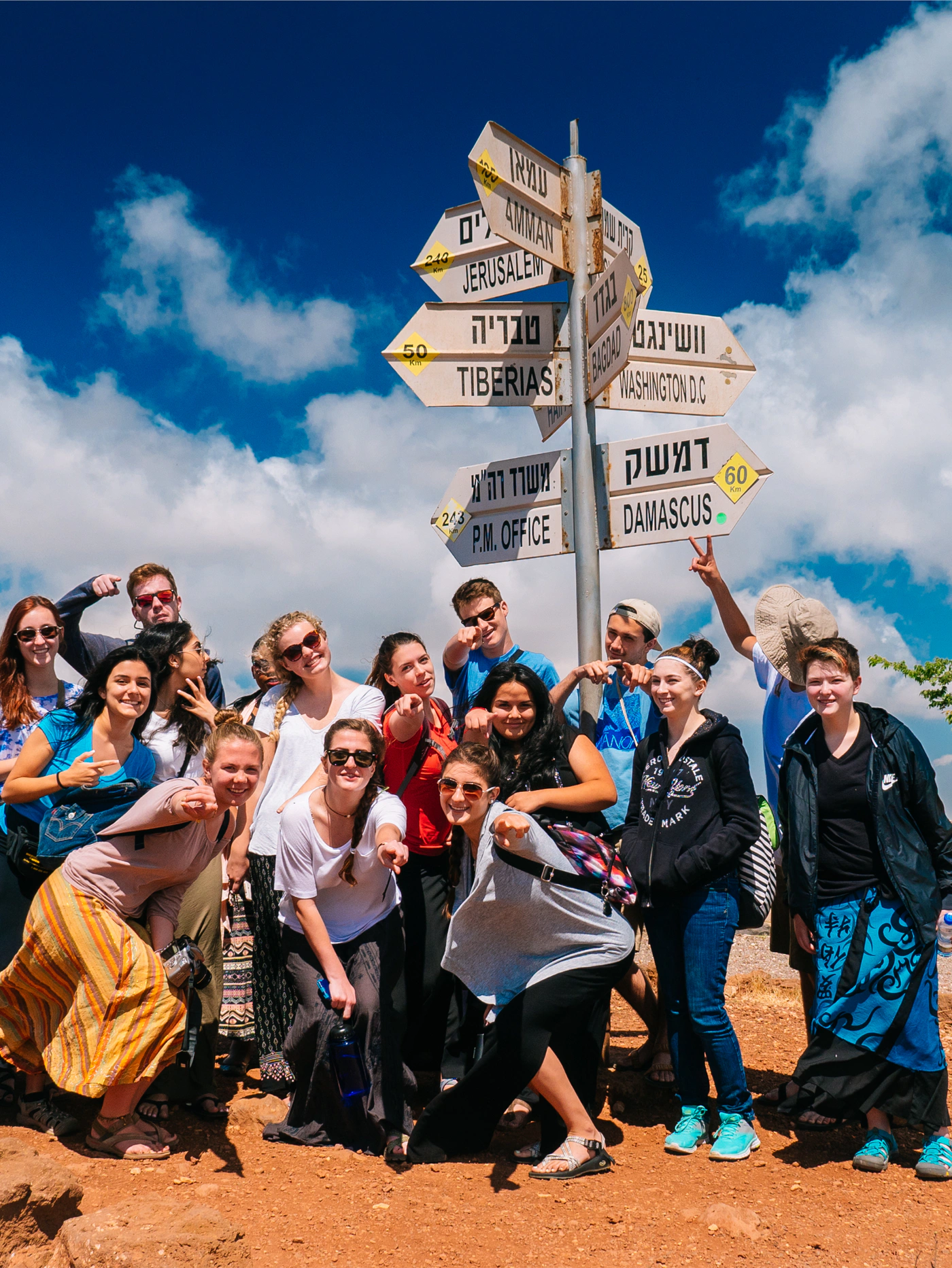 Students posing and pointing beneath a signpost to Jerusalem, Tiberias, Amman and Damascus under a blue sky