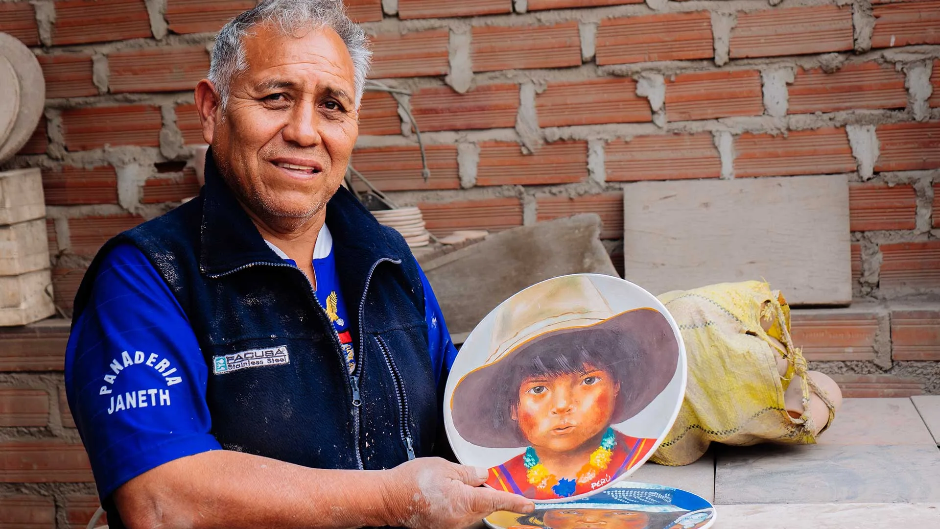 Cover of Meet the Artisans: Jose Yaksa: Jose Yaksa holds a hand-painted ceramic plate of a child in a hat, by a brick wall
