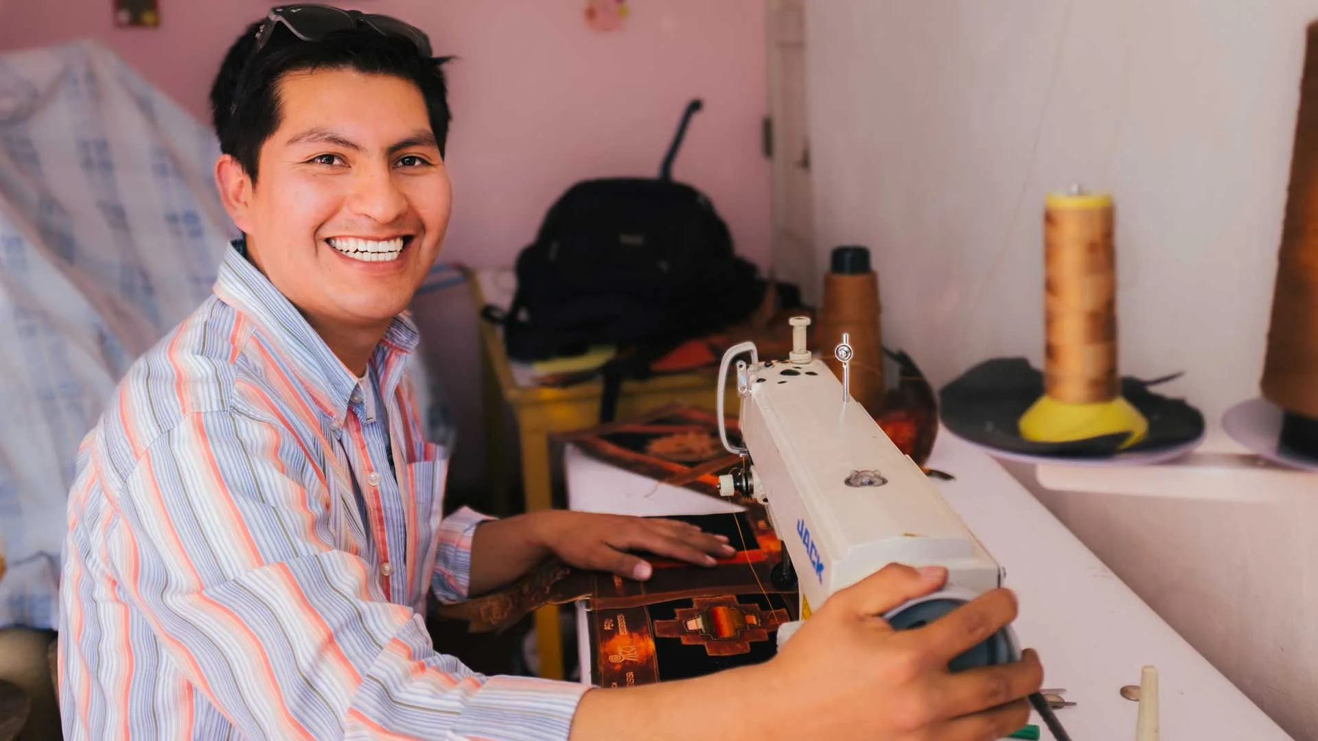 Cover of Meet the Artisans: Lenin: Lenin smiles at a sewing machine as he stitches tooled leather in a small pink room