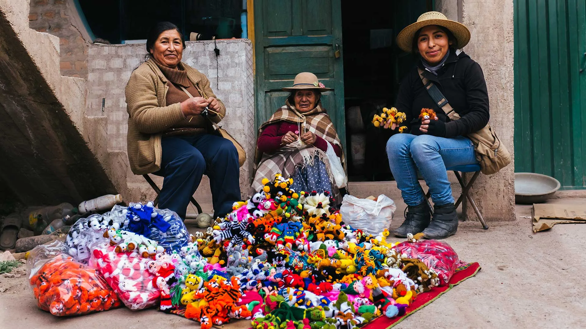 Cover of Meet the Artisans: Mabel: three women knit outside a doorway beside a pile of small knitted animal toys