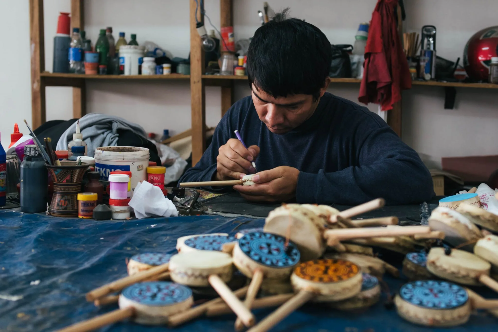 An artisan at a workbench painting a small gourd drum with a fine brush, a pile of painted drums in front of him and shelves of paint pots behind