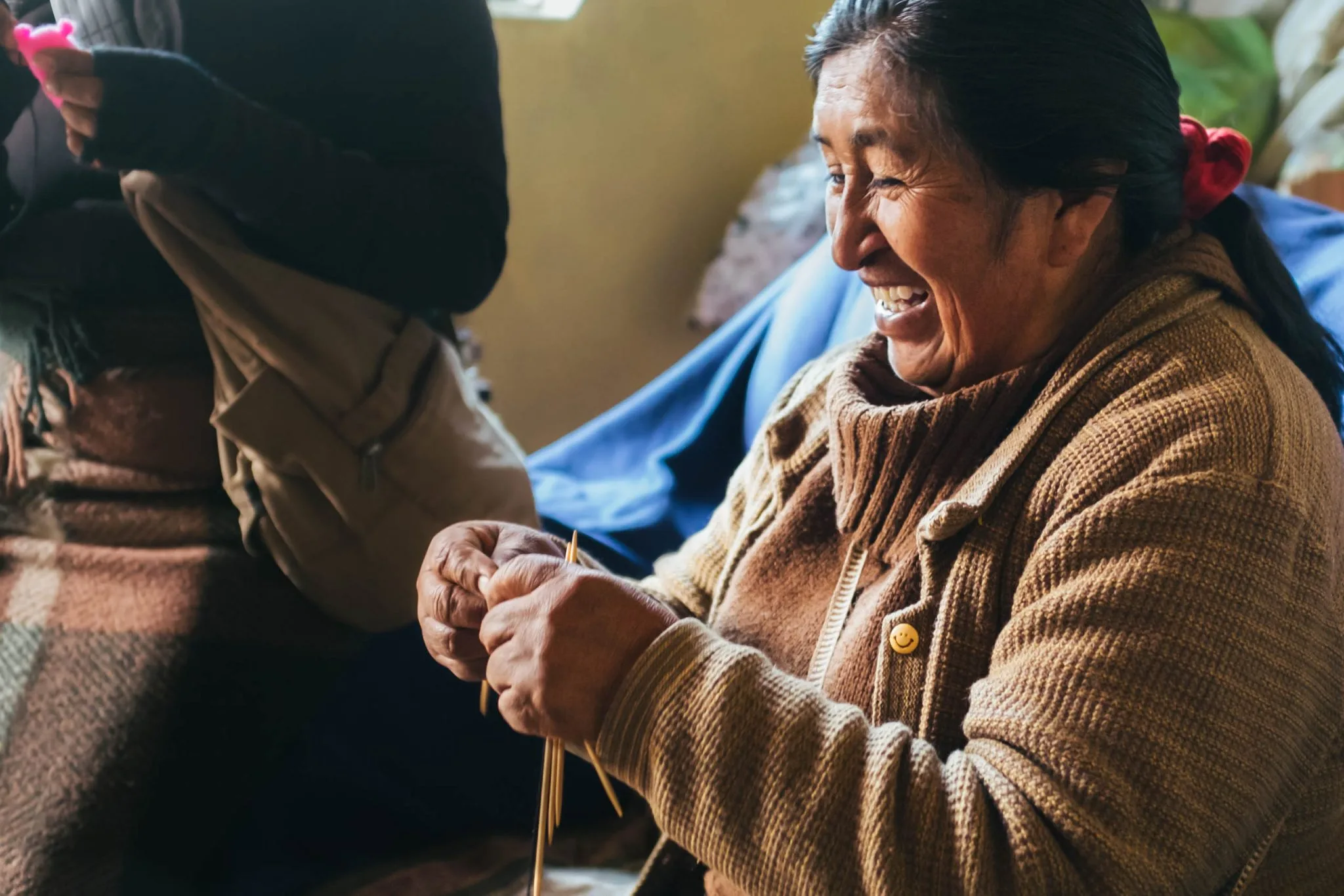 A woman in a brown knitted cardigan laughing as she knits