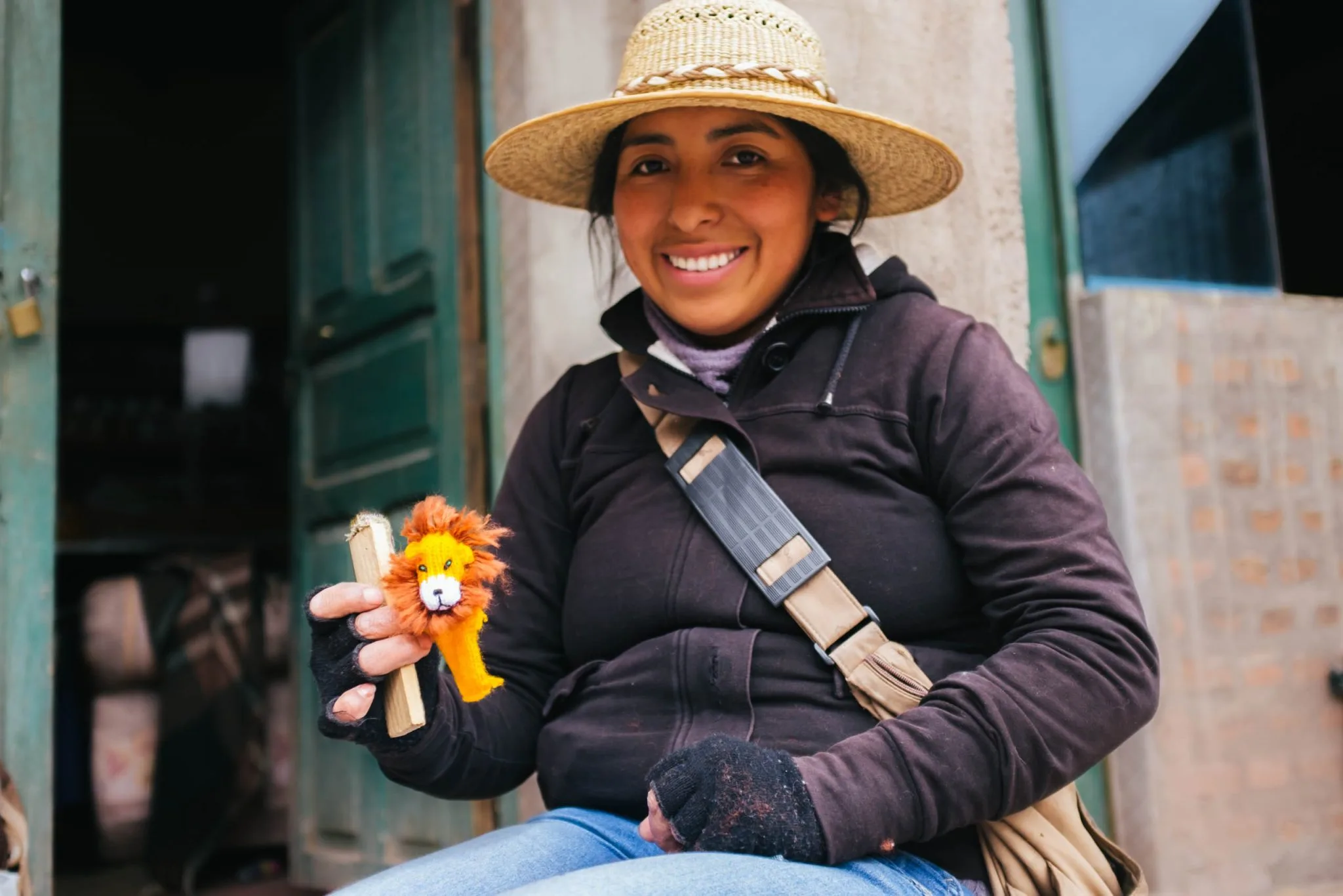 A smiling young woman in a straw hat sitting in a doorway, holding up a knitted orange lion finger puppet