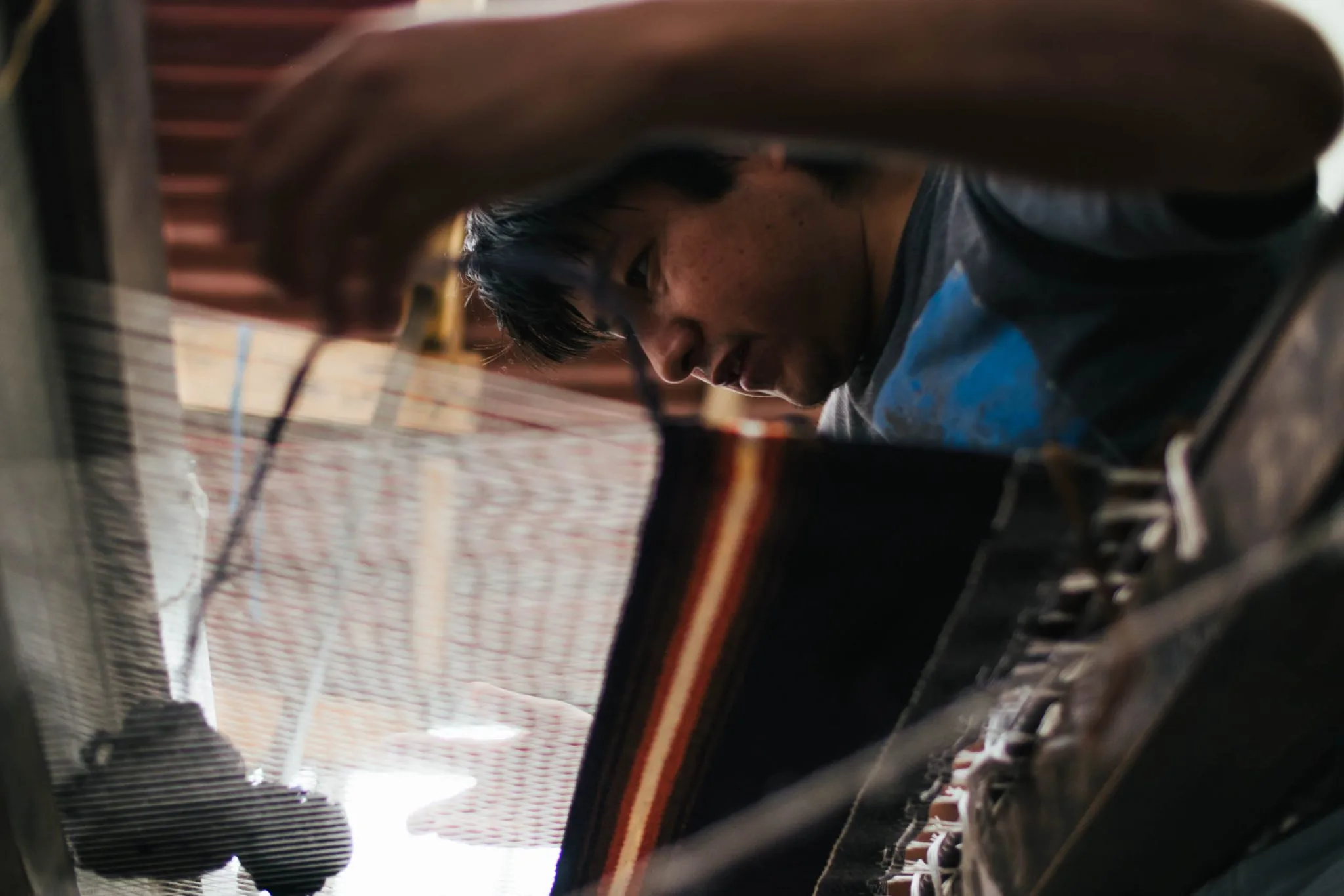 A weaver seen through the threads of a floor loom, a striped rug taking shape